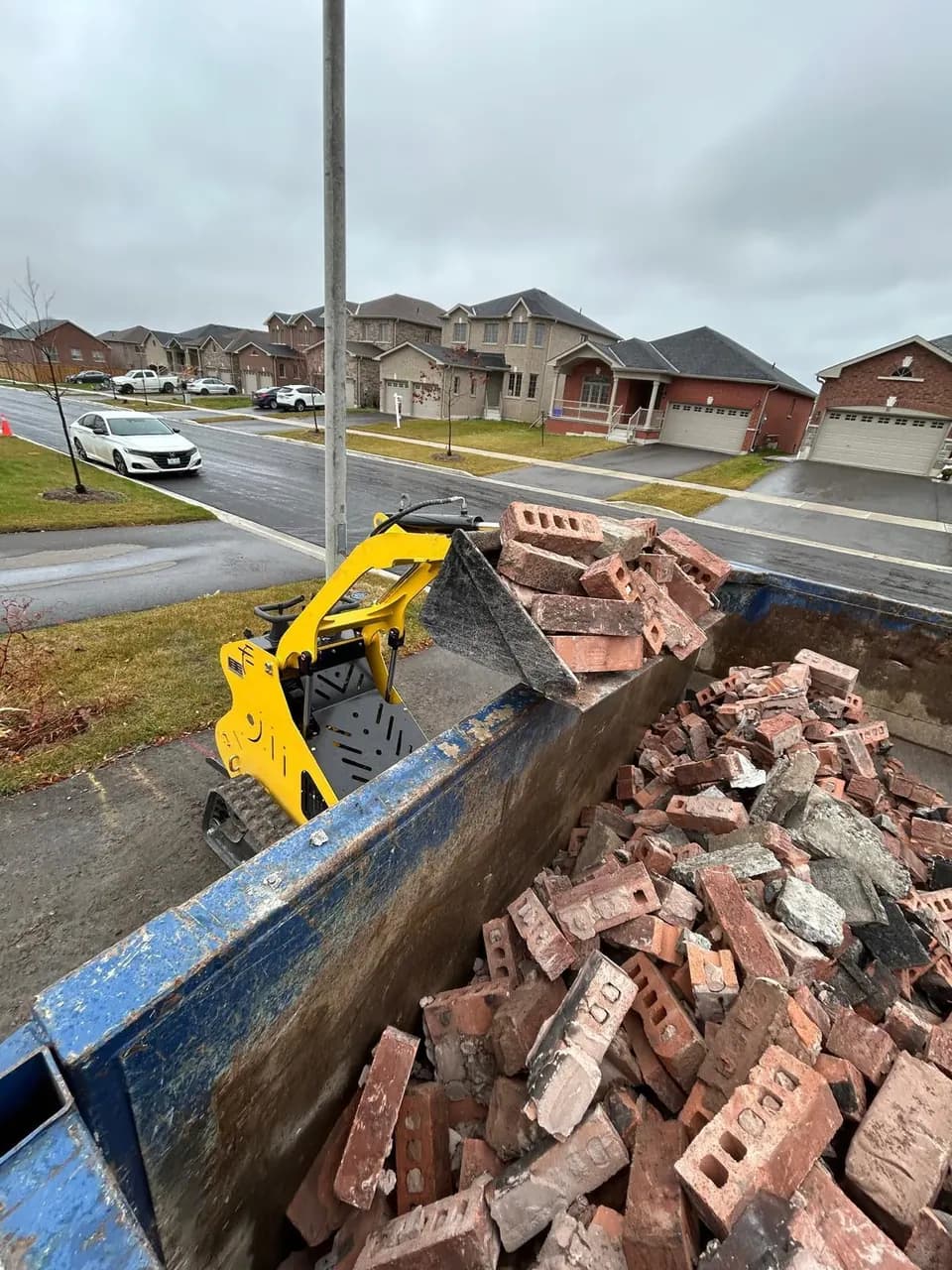 Our mini stand-on track loader loading bricks into a disposal bin on a residential GTA street — fits through a standard 36-inch gate.